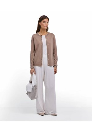 Femme portant un cardigan beige boutonné, un pantalon large blanc, tenant un sac fourre-tout à carreaux avec une écharpe blanche sur fond blanc.