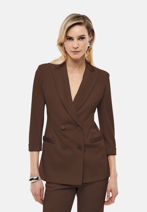 Donna con capelli biondi che indossa un blazer doppiopetto marrone e pantaloni abbinati, accessoriata con un braccialetto d'argento e orecchini geometrici.