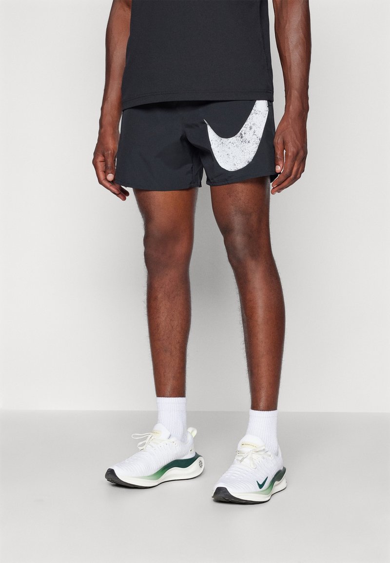 Mustat urheilushortsit, joissa on suuri valkoinen Nike-merkki, yhdistettyinä valkoisiin ja vihreisiin juoksukenkiin sekä valkoisiin nilkkasukkaihin.
