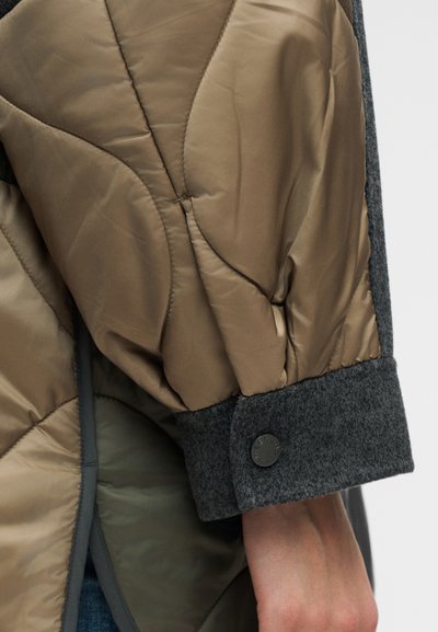 Μανίκι καπιτονέ καφέ μπουφάν με γκρι μάλλινη μανσέτα και μαύρο κουμπί, που δείχνει τον καρπό και μέρος του πουλόβερ από μέσα.