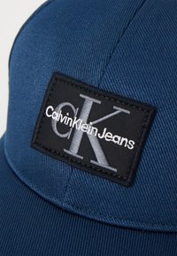 Tumesinine müts, mis on valmistatud tekstuurist kangast, millel on must ristkülikukujuline kleebis valge "CalvinKleinJeans" logo ja hõbedased "C.K." initsiaalid.