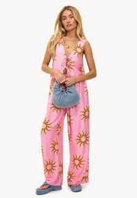 Mono sin mangas de color rosa con un motivo de sol en marrón, atado en la parte delantera. Combinado con un bolso de mano azul y sandalias de plataforma azules.