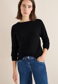 Maglione a coste nero con maniche a tre quarti, dotato di scollatura ampia. Abbinato a jeans in denim blu, che enfatizzano uno stile casual.