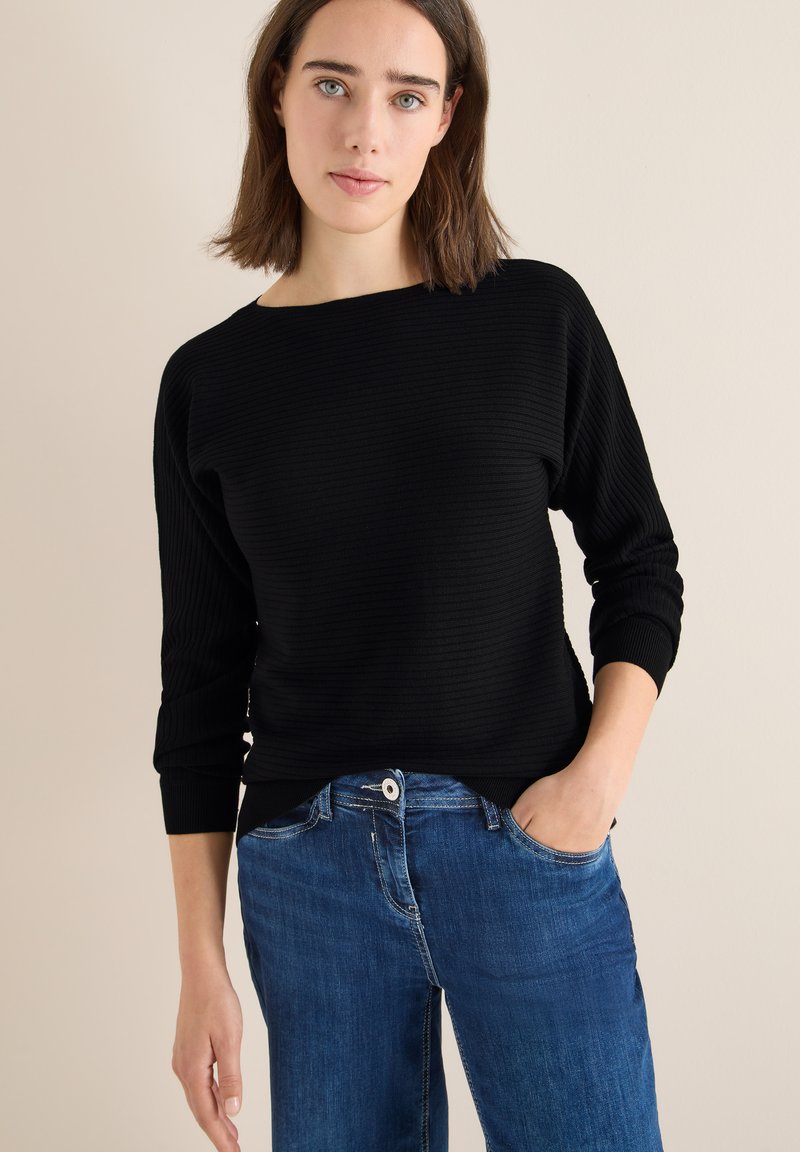 Maglione a coste nero con maniche a tre quarti, dotato di scollatura ampia. Abbinato a jeans in denim blu, che enfatizzano uno stile casual.