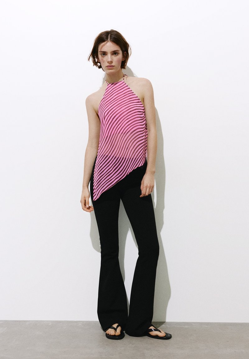 Femme debout contre un mur blanc portant un top dos nu rayé rose, un pantalon évasé noir et des sandales noires.