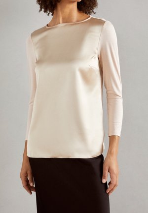 Woman wearing a cream satin sleeveless blouse over a light pink long-sleeve top and a black skirt, standing against a plain background.