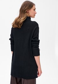 Kobieta z włosami do ramion, w czarnym golfie i kolczykach w kształcie kół, stoi bokiem na jednolitym tle.