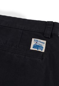 Czarne spodnie z kanwy z prostokątną, tkana metką z niebieskim tekstem i grafiką ciężarówki. Zawierają pasek i kieszeń z tyłu.