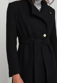 Abrigo negro de mezcla de lana con cintura ceñida por un cinturón, diseño de doble botonadura, acentos de botones dorados y un alto cuello. Textura suave y corte entallado.