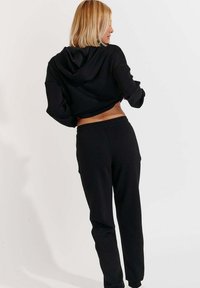 Schwarzer, kurzer Hoodie mit Kapuze, kombiniert mit schwarzen Jogginghosen. Der Stoff wirkt weich und glatt und hat einen lässigen Schnitt.