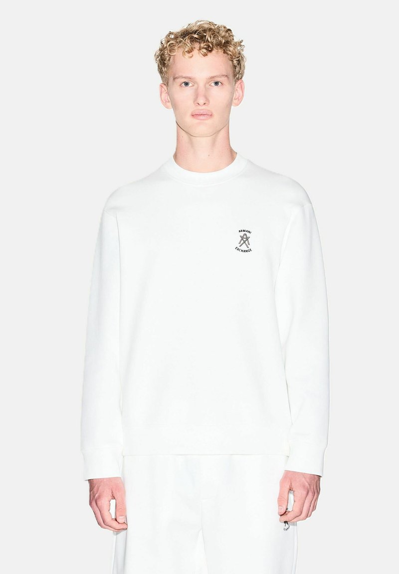 Jeune homme aux cheveux bouclés blonds portant une chemise blanche à manches longues et un pantalon blanc assorti avec un petit logo Armani Exchange.