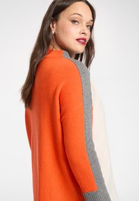 Der farbblockierte Pullover zeigt orange-, grau- und cremefarbene Abschnitte mit einem Rippbündchen-Design. Der Stoff hat eine weiche Textur.
