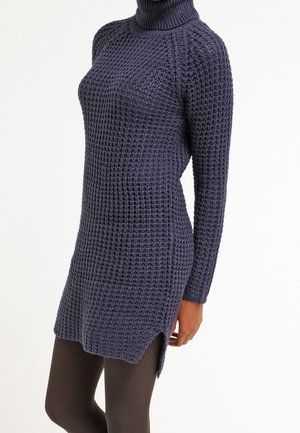 Femme portant une robe pull à col roulé tricotée bleu foncé avec texture gaufrée et fentes latérales, associée à des leggings foncés.