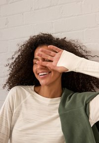 Light beige long-sleeve top with subtle stripes, paired with a dark green garment draped over the shoulder. Textured fabric, relaxed fit.
