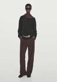 Maglione nero con un accento in pelliccia marrone, pantaloni a gamba larga in suede marrone e una grande cintura rotonda dorata. Occhiali da sole neri completano il look.