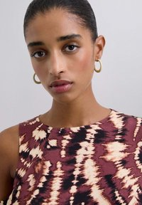 Una joven con el cabello liso recogido lleva pendientes de aro dorados y una blusa sin mangas con un patrón abstracto en púrpura, negro y crema.