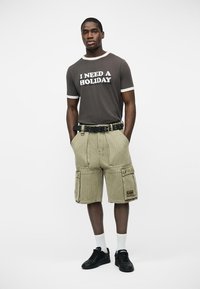 Mężczyzna stoi z rękami w kieszeniach, ubrany w czarną koszulkę z napisem "POTRZEBUJĘ WAKACJI", beżowe spodnie typu cargo, białe skarpetki i czarne trampki na białym tle.