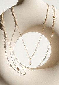 Collane a più strati di tono dorato con catene testurizzate, caratterizzate da molteplici perle e un pendente centrale a forma di goccia, esposte su uno sfondo neutro.