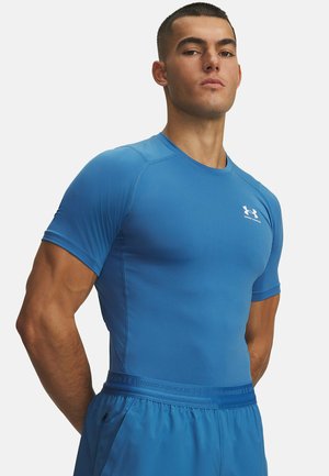 Muscular man with short hair wears tight blue Under Armour athletic shirt and matching blue shorts, standing with arms behind back.