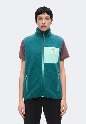 Persona joven con cabello corto que lleva un chaleco de forro polar azul verdoso con bolsillo en el pecho color verde menta y cremallera frontal sobre una camisa marrón de manga corta.
