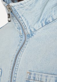 Ljusblå denimjacka med hög krage, silverdragkedja och sidofickor med sömnadsdetaljer. Tydligt texturmönster.