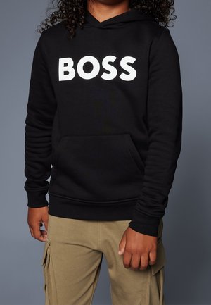 Persoon draagt een zwarte hoodie met "BOSS" in witte letters en beige cargobroek tegen een effen grijze achtergrond.