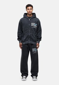 Zwarte zip-up hoodie met voorzakken en witte tekst; bijpassende joggingbroek met vergelijkbare tekst. Beide hebben verfspetters als accent.