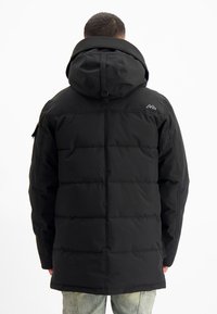 Man in een zwarte gewatteerde winterjas met capuchon, gezien vanuit de rug, staand tegen een effen witte achtergrond.