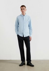 Jasnoniebieska koszula z długim rękawem zapinana na guziki, w połączeniu z ciemnoszarymi spodniami i czarnymi butami. Koszula ma małe logo na klatce piersiowej.