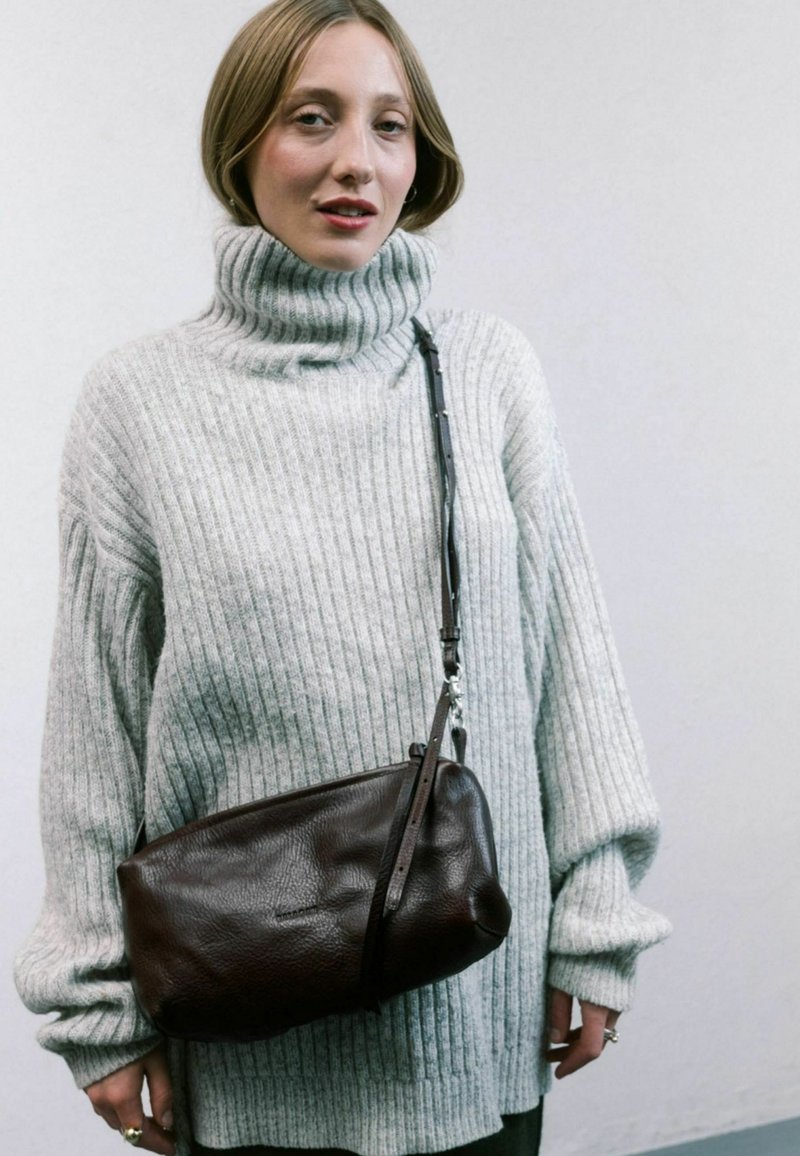 Maglione grigio a coste con colletto a lupetto, vestibilità ampia; borsa a tracolla in pelle marrone con finitura testurizzata e tracolla regolabile. Sfondo uniforme.