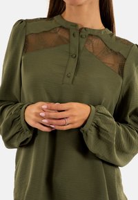 Blusa verde oliva con tejido texturizado, tapeta de botones y detalles de malla translúcida en los hombros y en el escote. Mangas largas y abullonadas.