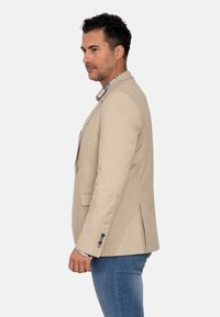 Blazer beige a medida con un corte estructurado, cierre de un solo botón y costuras detalladas, llevado sobre una camisa de cuadros.