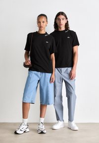 To modeller som har på seg svarte Lacoste t-skjorter; en i lys blå denimshorts og sneakers, den andre i lyse stripete bukser og hvite sko.