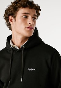 Μαύρη κουκούλα με κορδόνι, διαθέτει λευκό ραμμένο λογότυπο. Κάτω από αυτήν βρίσκεται ένα ανοιχτό καρό πουκάμισο με γιακά, παρουσιάζοντας στρωματοποιημένες υφές.