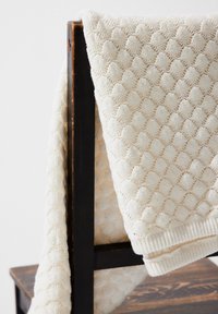 Strickdecke in Creme mit einem strukturierten, erhabenen Muster und gerippten Rändern, über einen dunkelbraunen Stuhl drapiert.