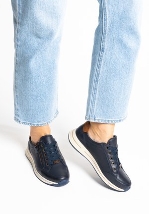 Persona con jeans azzurri chiari e sneaker in pelle blu navy con suole bianche, toppa marrone sul tallone e dettaglio con cerniera laterale.