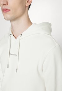 White hoodie with a kangaroo pocket, adjustable drawstrings, and a printed label "CASUAL" on the chest. Cotton blend material, smooth texture.
