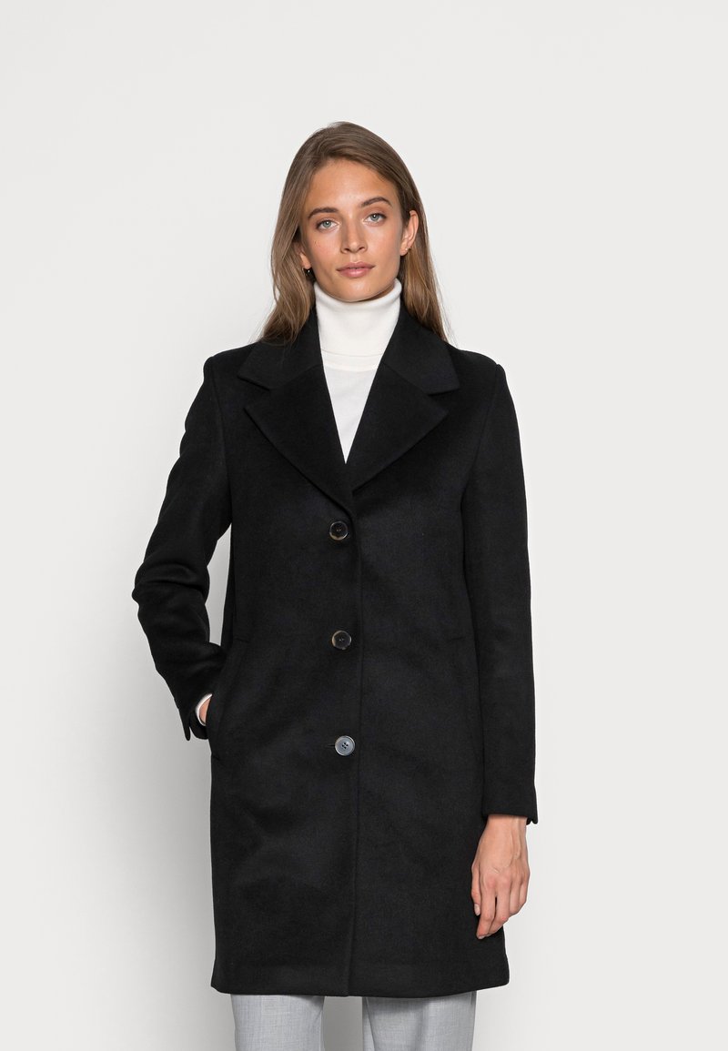 Melnā vilnas mētelis ar ieliektu apkakli, trim podziņām un sānu kabatām. Gluda tekstūra, līdz ceļiem garš dizains, valkāts virs baltas turtleneck kreklas.