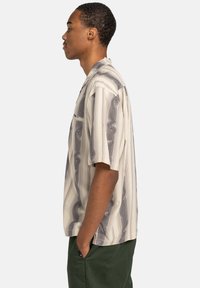 Short-sleeve button-up shirt in light beige with abstract gray wave patterns, featuring a relaxed fit and a single chest pocket.