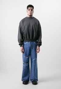 Sweatshirt gris foncé avec des poignets côtelés, associé à un jean bleu clair déchiré avec des déchirures et des éclaboussures de peinture. Baskets noires aux pieds.