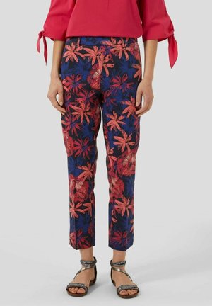 Vrouw die cropped bloemenbroek draagt in rood, blauw en zwart, gecombineerd met een rode blouse en zilveren sandaaltjes met bandjes op een effen achtergrond.