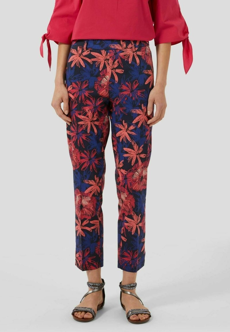 Femme portant un pantalon court à fleurs rouge, bleu et noir associé à un chemisier rouge et des sandales argentées à brides sur un fond uni.