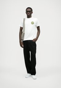 Balts T-krekls ar apaļu dzeltenu un zaļu grafiku, apvienots ar brīviem melniem biksēm un baltām sporta apaviem, papildināts ar bēša plecu somu.