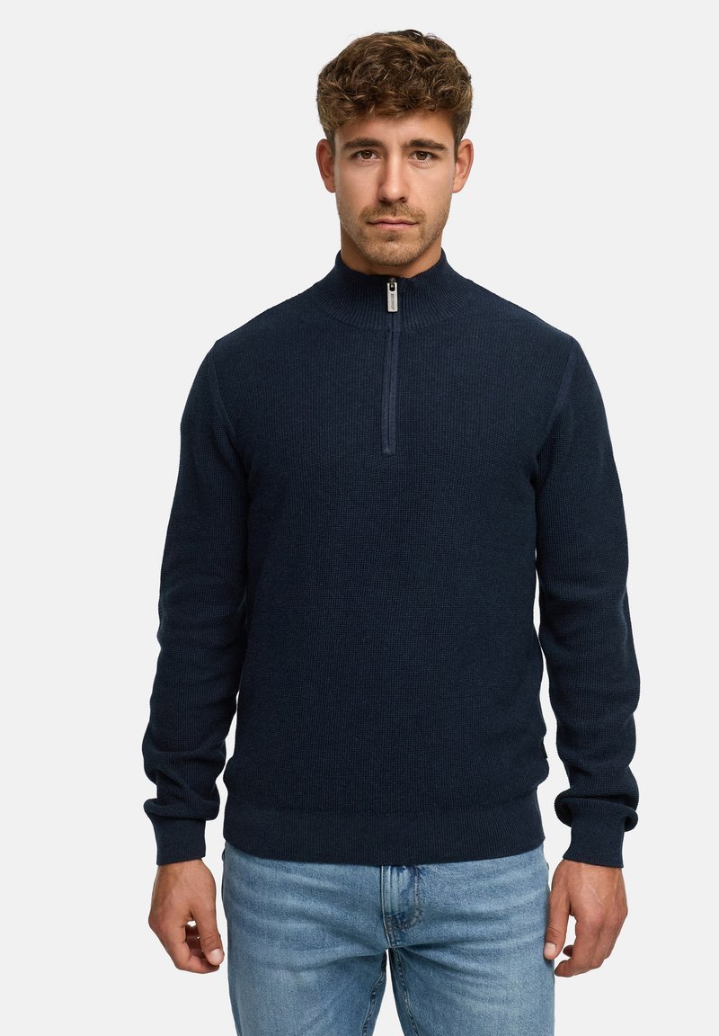 Marinefarbener Pullover mit Viertelreißverschluss aus strukturiertem Strickstoff, mit Stehkragen und gerippten Bündchen, kombiniert mit hellblauen Jeans.