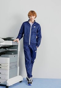 Σαλοπέτα και παντελόνι ναυτικού χρώματος, φτιαγμένα από ελαφρύ υλικό, με πλήρες φερμουάρ, άσπρες λεπτομέρειες και λογότυπο. Τοποθετημένα δίπλα σε έναν φωτοτυπικό μηχάνημα.