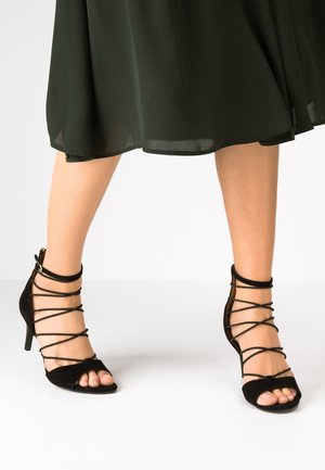 Schwarze Wildleder-Sanduhr mit offenem Zehenbereich und schmalen, überkreuzten Knöchelriemen. Glatte Textur und ein schlanker Absatz.