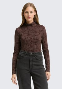 Braunes Langarmoberteil mit hohem Kragen und schwarzem Punktmuster, kombiniert mit hochgeschnittenen schwarzen Jeans mit normalen Taschen.