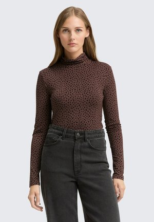 Braunes Langarmoberteil mit hohem Kragen und schwarzem Punktmuster, kombiniert mit hochgeschnittenen schwarzen Jeans mit normalen Taschen.