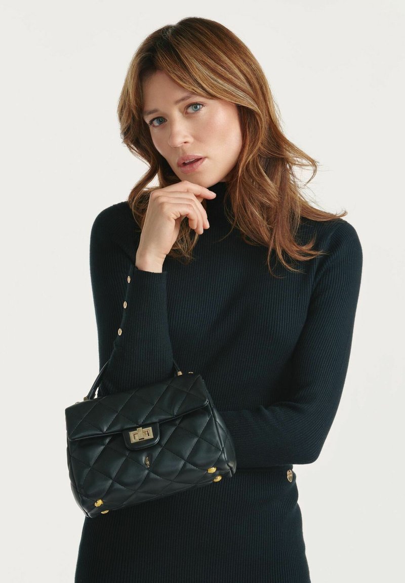 Kwiltowana czarna torba skórzana z złotymi akcentami, prostokątny kształt i uchwyt u góry. Tło w neutralnym kolorze, które podkreśla design torby.