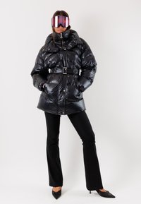 Giacca piumino nera, materiale lucido, design trapuntato, vita con cintura, colletto alto, abbinata a occhiali da sci rosa specchiati oversized, pantaloni neri, tacchi a punta.